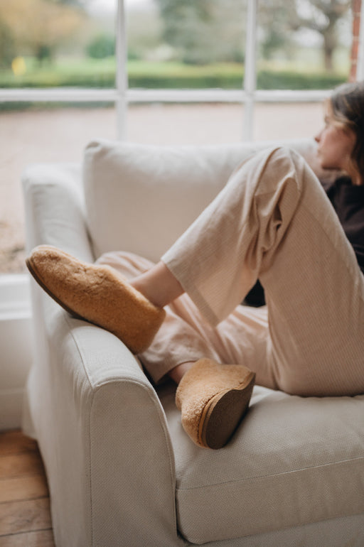 Side view of a camel coloured curly merino sheepskin boot slipper worn by a lady relaxing on a white sofa chair.
