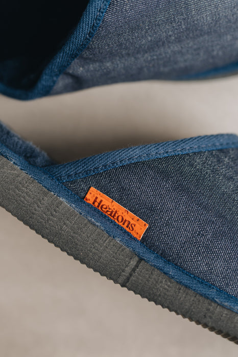 Close-up of navy,  denim covered, sheepskin mule slippers with a cork orange 'Heaton's' brand label displayed