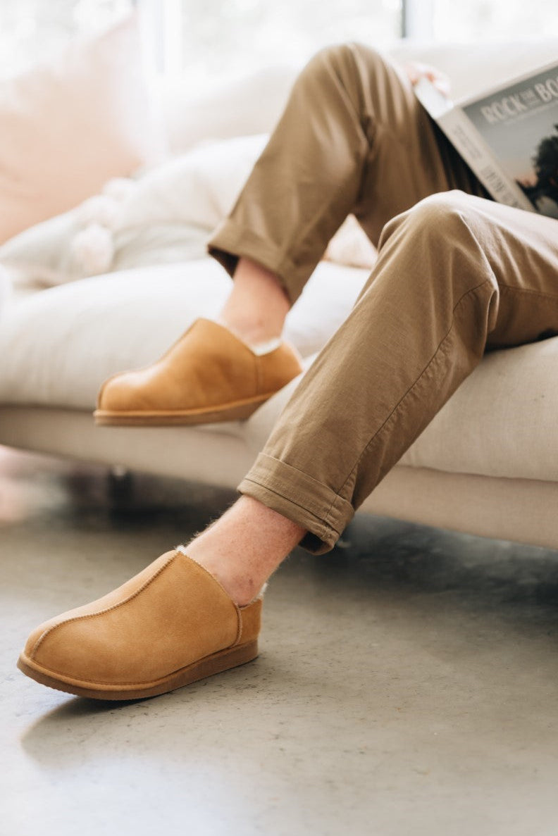Man sitting on a couch wearing tan Cadi Merino sheepskin slippers and reading a magazine.