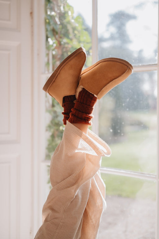 Tan sheepskin slippers on feet with rust coloured socks, in front of a window with a view of greenery.