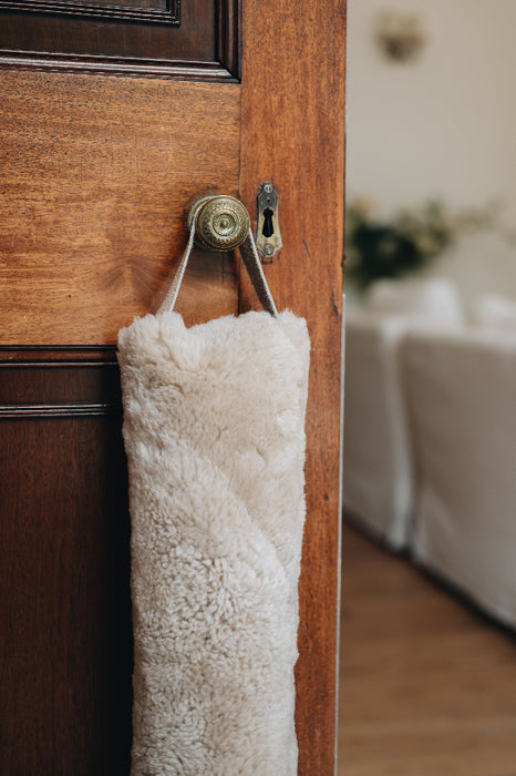 Oatmeal sheepskin draught excluder with leather handle hanging on a wooden door with a brass doorknob and keyhole
