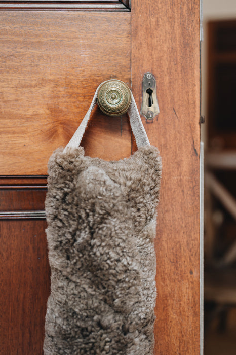 Wheat brown sheepskin draught excluder with leather handle hanging on a wooden door with a brass doorknob and keyhole.