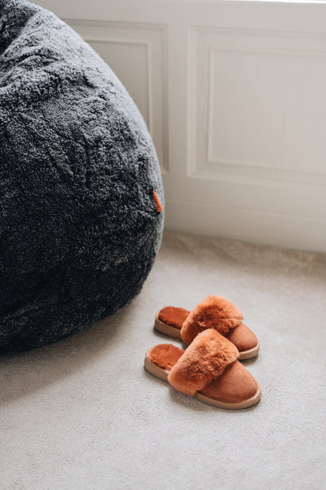Pair of rust coloured sheepskin slippers on a light  carpet next to a large dark sheepskin bean bag chair.