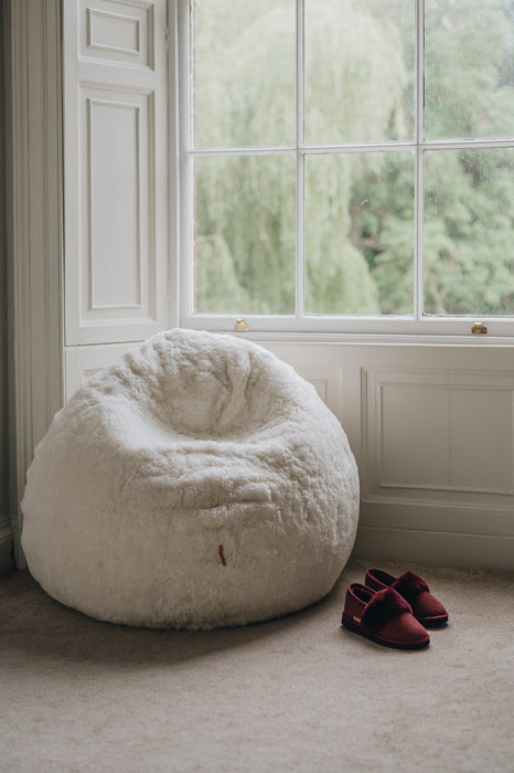 White sheepskin bean bag chair next to a window with a pair of slippers on a carpeted floor.