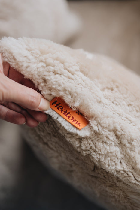 Close-up of an oat coloured sheepskin cushion with an orange  'Heatons' label, held by a hand.