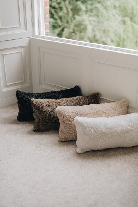 Four rectangular sheepskin cushions in black, brown, beige, and white on a carpeted floor near a window.