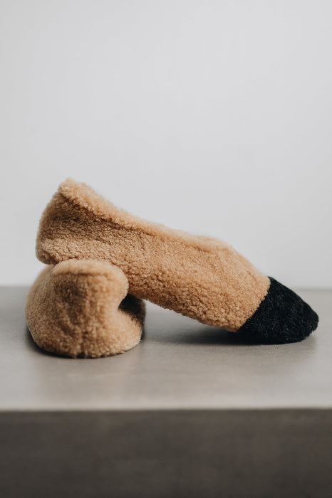 Pair of curly sheepskin tan slippers with black toes on a light grey surface