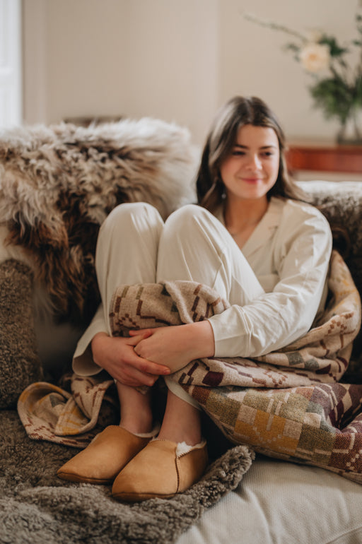 Grace soft soled slipper worn by a young woman with their feet up on a couch, surrounded by blankets and sheepskin rugs looking cosy.