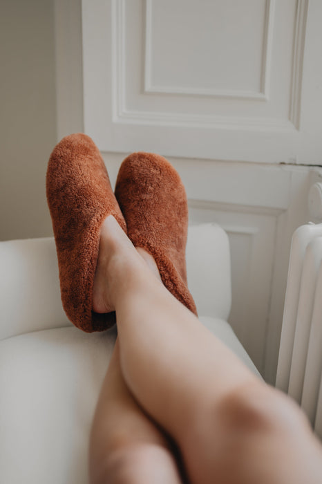 Ladies feet up wearing a pair of curly merino sheepskin slip on slipper with curls on the outside and inside in a rich, warm rust colour.