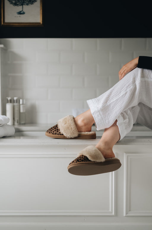 Women sat on edge of bath wearing leopard printed merino sheepskin slippers called Veronica.