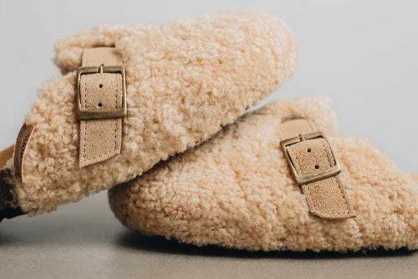 Side view of a vanilla coloured curly sheepskin mule, showing of it's adjustable sheepskin strap and quality buckle