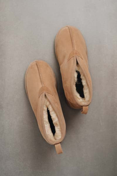 A pair of tan slip-on sheepskin slippers with white shearling lining, photographed from a top-down angle on a textured grey background. Slipper is named Erin.