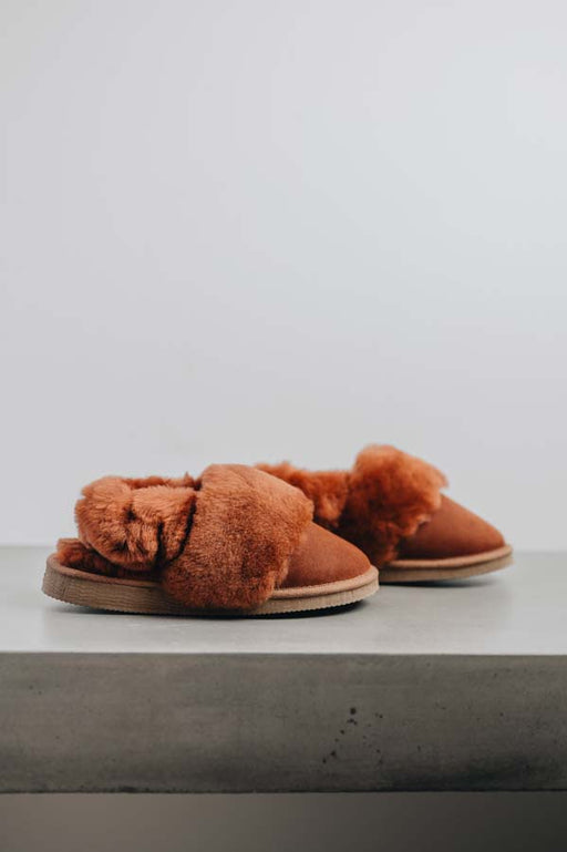 Pair of rust coloured sheepskin slippers with fluffy wool cuff  and elasticated heel band on a light surface