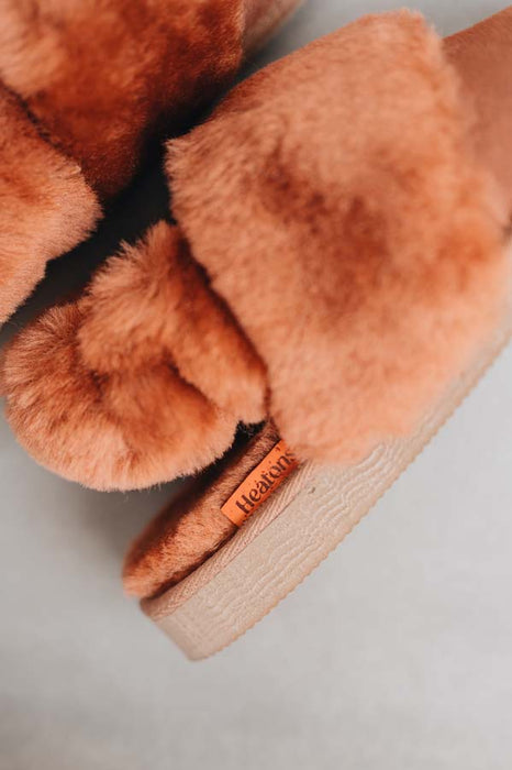 Close-up of a pair of fluffy rust coloured sheepskin slippers with an elasticated band and an orange cork Heatons brand label on a light background