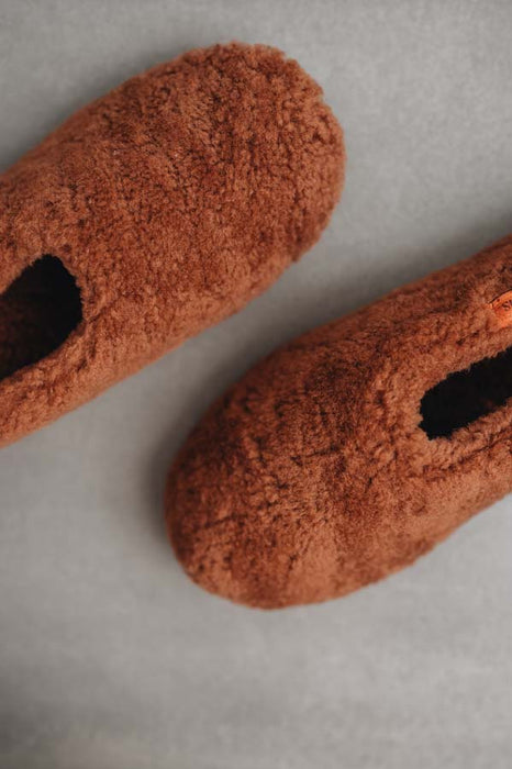 Pair of rust coloured curly sheepskin slippers on a light grey background