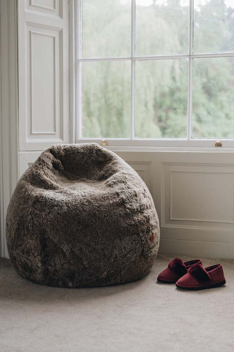 A large, plush wheat coloured sheepskin bean bag chair sits on a light carpeted floor next to a pair of burgundy slip-on Margot slippers in front of a white window.