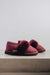 A pair of burgundy slip-on slippers with plush sheepskin cuff across the top, resting on a light grey concrete ledge against a neutral light grey background. A small tan Heatons brand tag is visible on the side of the front slipper.
