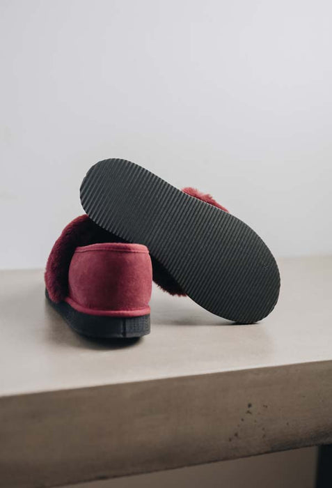 A pair of Bordeaux coloured slip-on slippers with one shoe turned upside down to show the textured black sole, resting on a grey stone surface