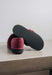 A pair of Bordeaux coloured slip-on slippers with one shoe turned upside down to show the textured black sole, resting on a grey stone surface