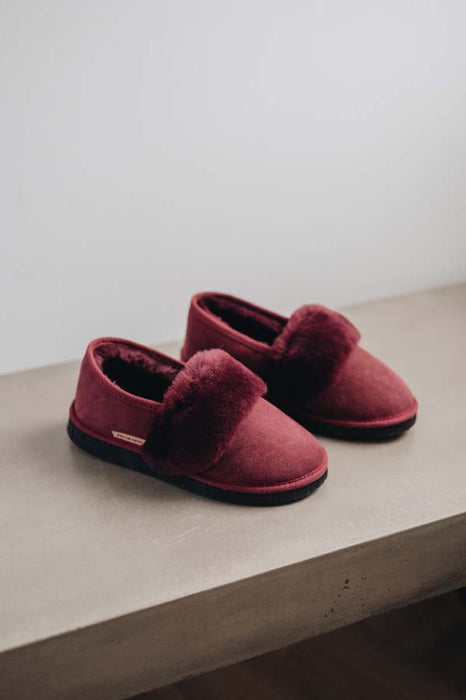 A pair of burgundy slip-on slippers with plush sheepskin cuff and black soles, displayed on a light grey concrete ledge against a neutral off-white wall.