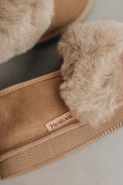 Close-up of a pair of taupe sheepskin slippers with fluffy cuff, featuring a small tan cork 'Heaton's' brand tag on the side.