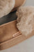 Close-up of a pair of taupe sheepskin slippers with fluffy cuff, featuring a small tan cork 'Heaton's' brand tag on the side.