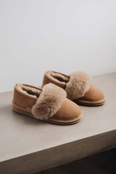 A pair of taupe coloured sheepskin slippers with fluffy shearling cuff, resting on a light grey concrete surface against a neutral off-white background.
