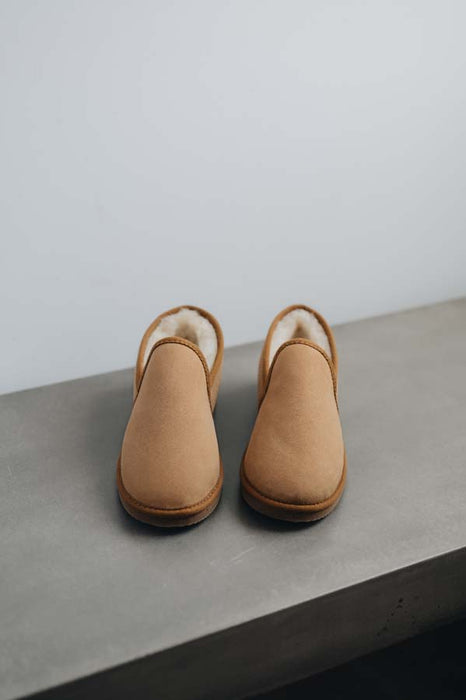 A pair of tan men's sheepskin slippers with a soft cream-colored shearling lining, displayed on a grey concrete bench