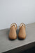 A pair of tan men's sheepskin slippers with a soft cream-colored shearling lining, displayed on a grey concrete bench