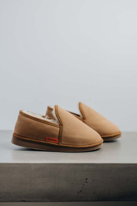 Reggie tan men's merino sheepskin slippers with a soft white fleece lining, displayed on a concrete ledge against a light background.