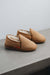 A pair of men's tan sheepskin slippers with a soft cream fleece lining, resting on a grey concrete ledge against a light gray wall.