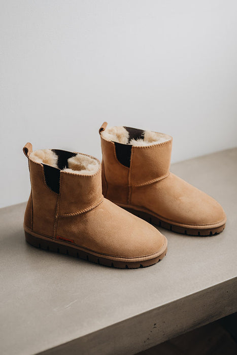 Pair of tan sheepskin chelsea style boots with dark brown elastic on a light surface.