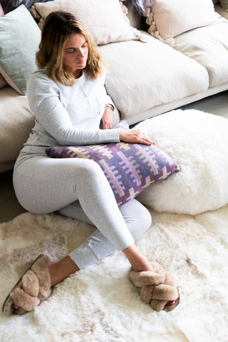 Wide view of Icelandic White Patchwork Sheepskin Melange Rug in a living room. Features white, beige and grey coloured patches. A woman sits on top of the rug with Gabriella beige slippers and holding a Melin Tregwynt purple patterned cushion.