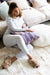 Wide view of Icelandic White Patchwork Sheepskin Melange Rug in a living room. Features white, beige and grey coloured patches. A woman sits on top of the rug with Gabriella beige slippers and holding a Melin Tregwynt purple patterned cushion.