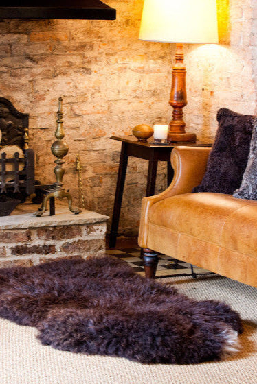 Cosy Brown British Sheepskin Rug, in front of the fire on the floor with accent Sheepskin cushions on a  leather sofa next to it. 