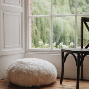 White sheepskin footstool on a wooden floor with a large window in the background showing greenery.