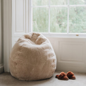 Oatmeal sheepskin bean bag chair next to a window with a pair of slippers on a carpeted floor.