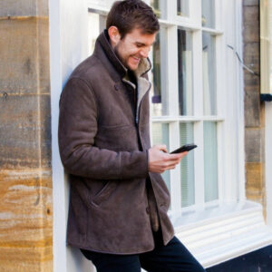 Men's Sheepskin Coat. Male model wears brown Sheepskin coat, with hand in pocket, looking down at phone.